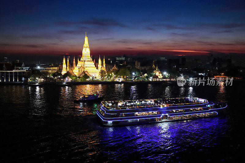 泰国曼谷，湄南河上，一艘晚餐游船经过黎明寺(Wat Arun)，灯光照亮的滨水区，黄昏时水面的倒影，聚焦于前景